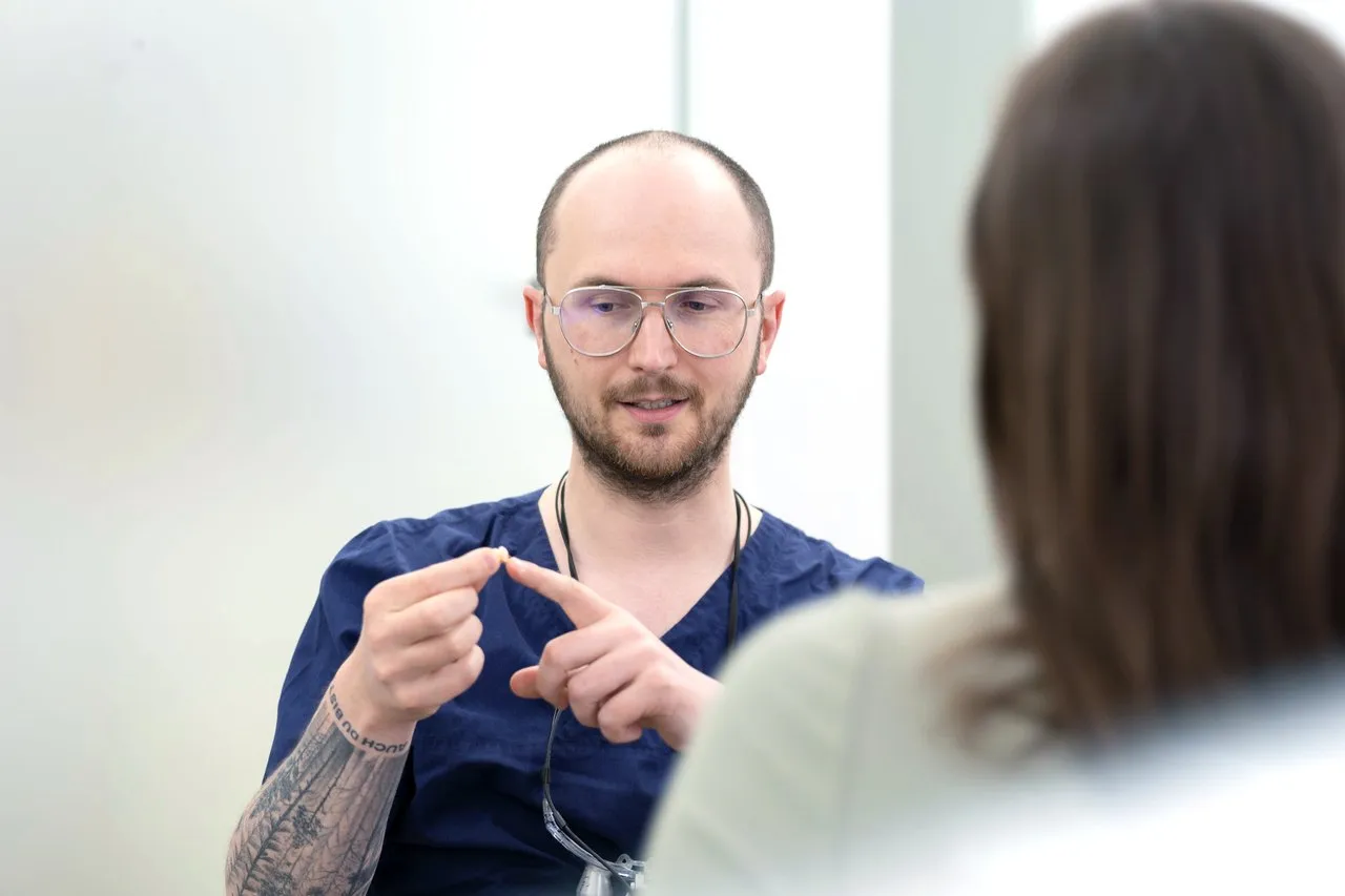 Zahnarzt im blauen Kasack erklärt einer Patientin im Behandlungszimmer etwas mit beiden Zeigefingern.