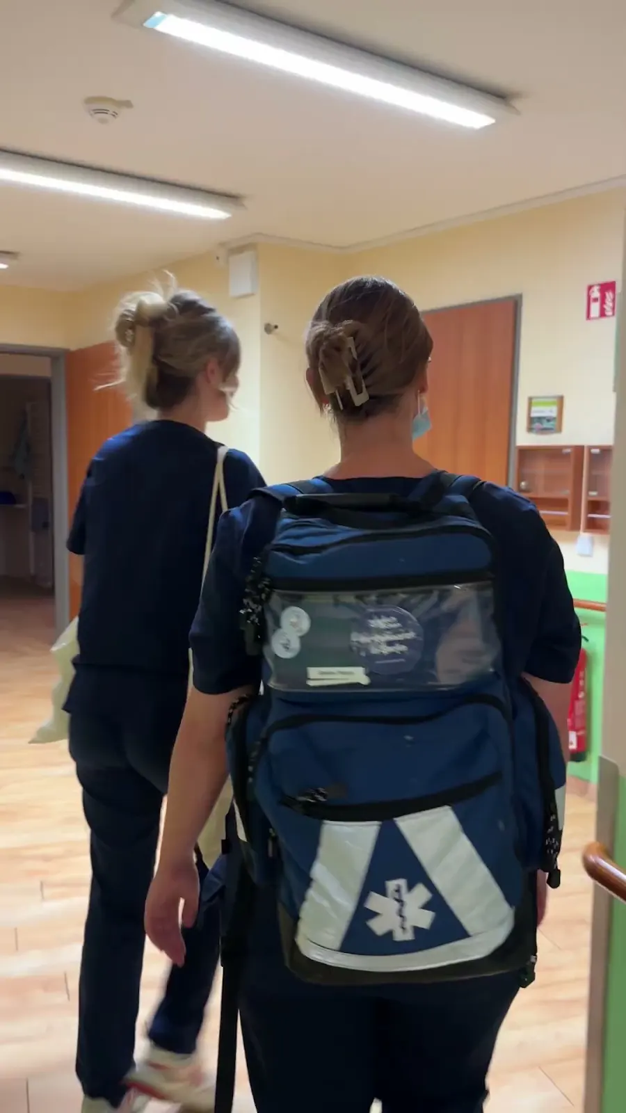 Original-Standbild aus dem VLOG: Zwei Teammitglieder transportieren mobile Ausstattung im Rucksack durch einen Heimflur.